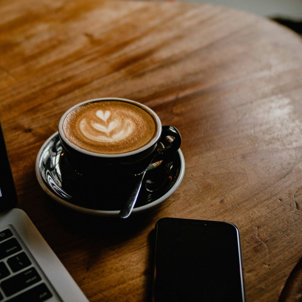 A latte on a wooden café table beside a laptop and phone